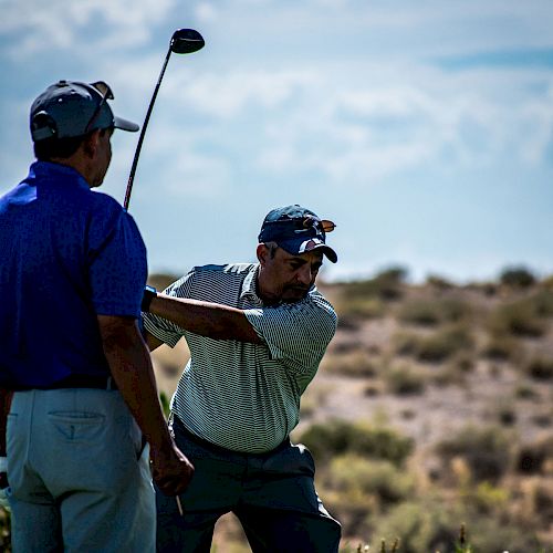 Two people are on a golf course; one swings a golf club while the other stands nearby watching. The background is a clear sky and landscape.