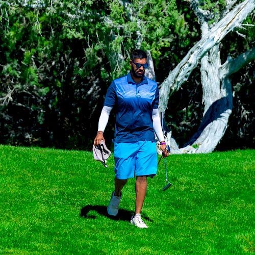 A person is walking on a golf course holding golf clubs and a towel, surrounded by green grass and trees in the background.