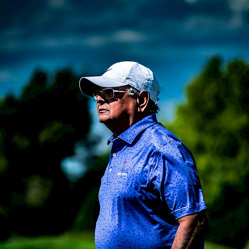 A person wearing a blue shirt and white cap stands outdoors, holding a golf club. The sky is partly cloudy, with trees in the background, ending the sentence.