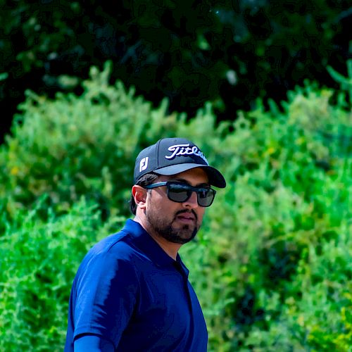 A person in a blue shirt and sunglasses, wearing a black baseball cap, stands in front of green foliage.