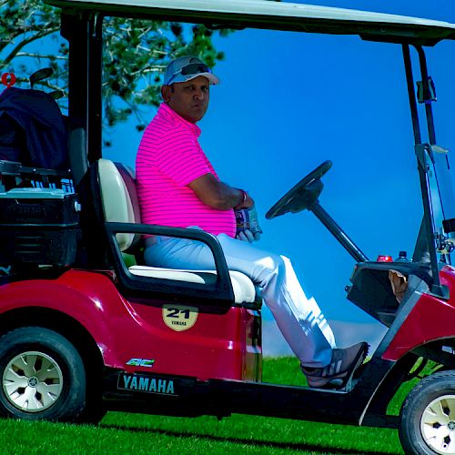 A person wearing a pink shirt and white pants sits in a red Yamaha golf cart on a grassy area, with trees and a blue sky in the background.