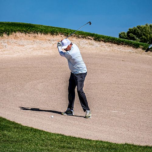 A golfer is making a shot from a sand trap on a golf course, surrounded by green grass and under a clear blue sky, with trees in the background.