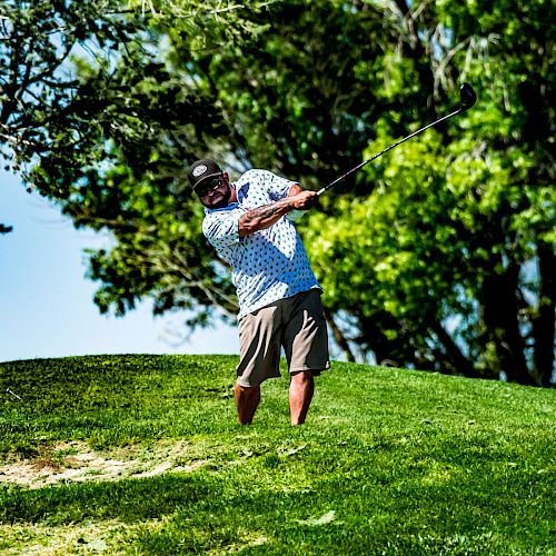 A person is playing golf, swinging a club on a green fairway, with trees in the background under a clear sky.