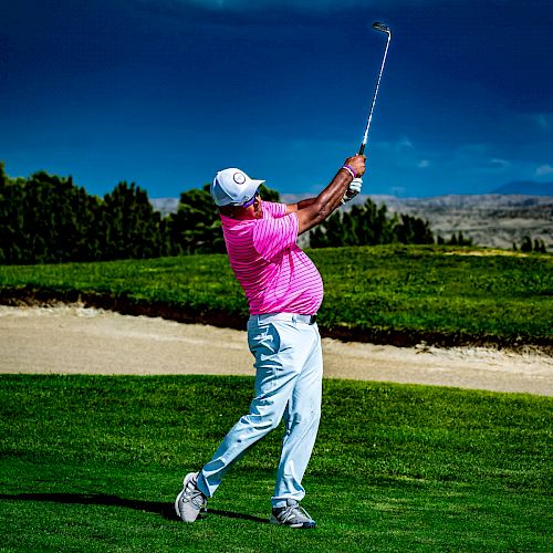 A golfer wearing a pink shirt and white pants swings his club on a green with a sand bunker and trees in the background under a cloudy sky.