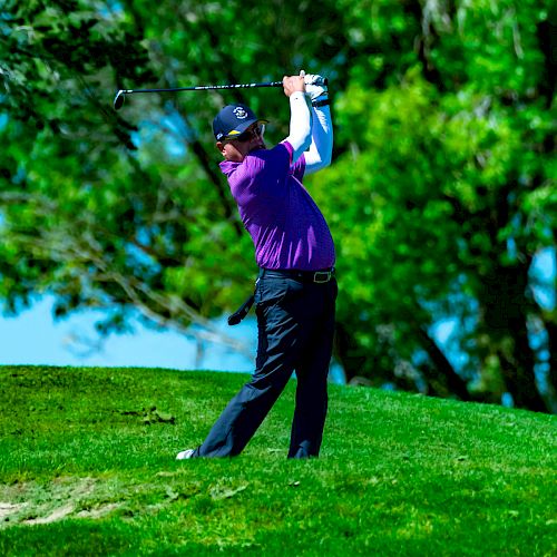 A person wearing purple and black golfing attire is captured mid-swing on a lush green golf course with trees in the background.