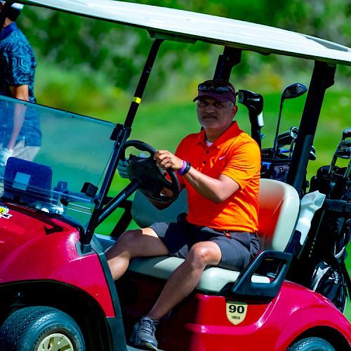 A person in an orange shirt is driving a red golf cart on a golf course, with golf clubs in the back, and another person is walking nearby at a distance.