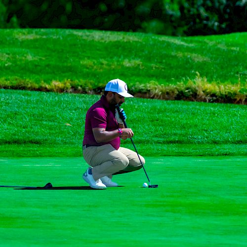 A person is kneeling on a golf course, focused intently on lining up a putt with a golf club in hand.