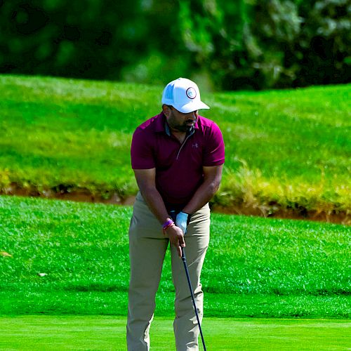 A person dressed in a maroon shirt and khaki pants prepares to swing a golf club on a lush green golf course.