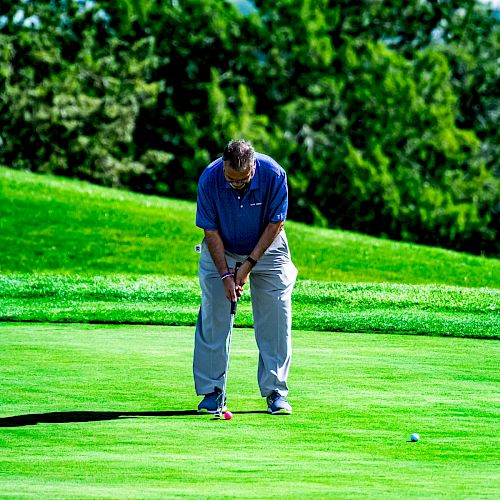 A person in a blue shirt and gray pants is putting a golf ball on a green golf course with trees in the background.