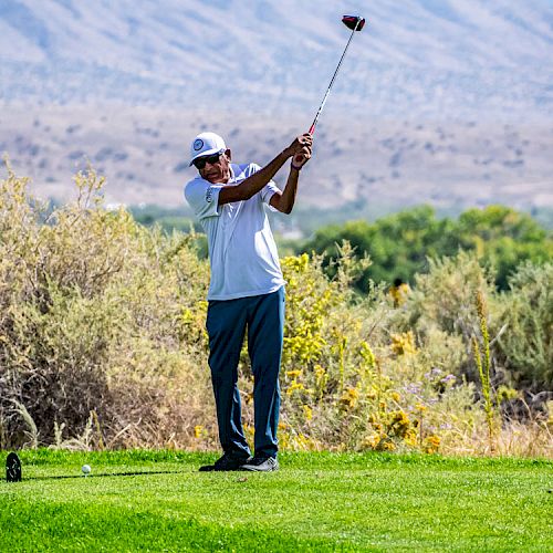 A person wearing a white cap and shirt swings a golf club on a green golf course against a backdrop of hills and trees.