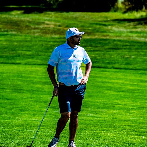 A person wearing a hat and casual sportswear is standing on a golf course, holding a golf club, and looking into the distance.