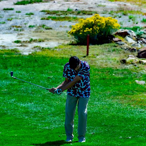 A person wearing a patterned shirt playing golf on a green course, swinging a club with a focused posture.