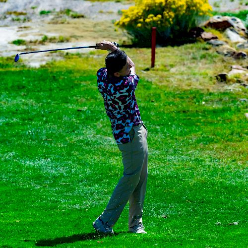 A person is playing golf, swinging a golf club on a green lawn with shrubs and rocky terrain in the background.