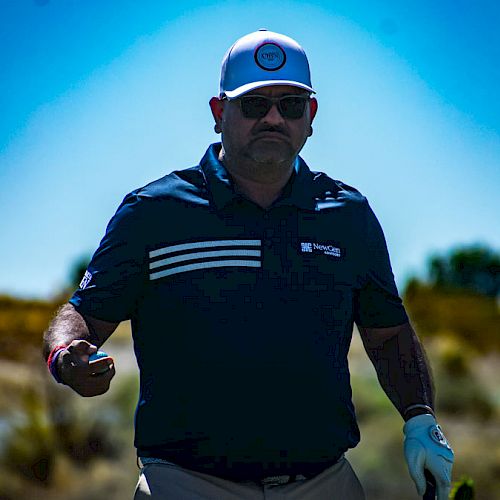 A man in sunglasses and a cap holds a golf ball while standing on a golf course under a clear blue sky.