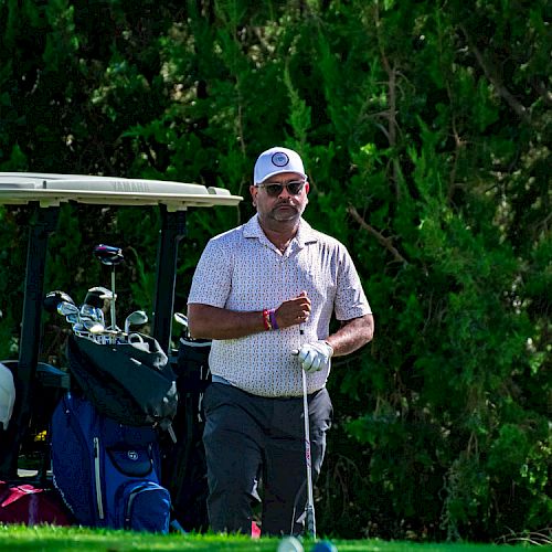 A person stands near a golf cart holding a golf club, with trees in the background and a sign visible on the right side.