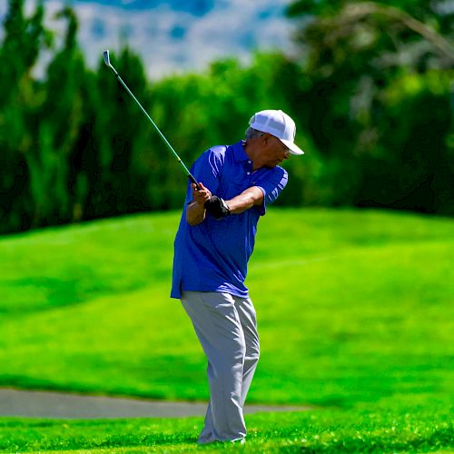 A person in a blue shirt and white hat is playing golf on a lush green course, swinging a club on a bright, sunny day.