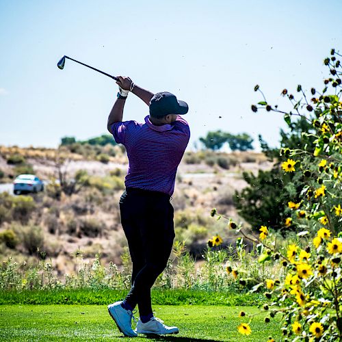 A person is mid-swing, playing golf on a lush green course under a clear sky, with sunflowers in the foreground and a car in the background.