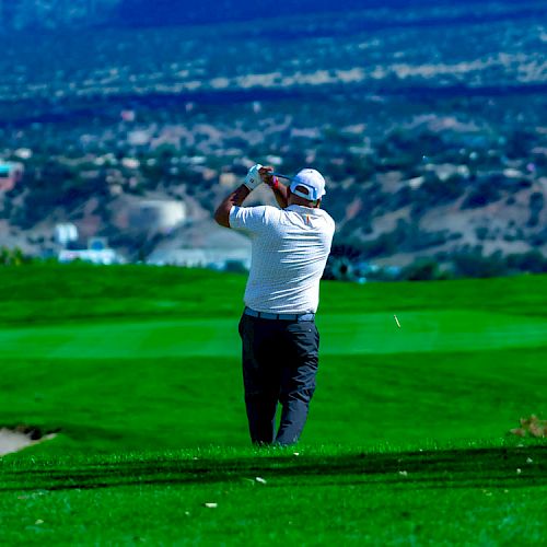 A person is playing golf on a green field with mountains and a valley in the background, wearing a white shirt and dark pants, swinging a club.