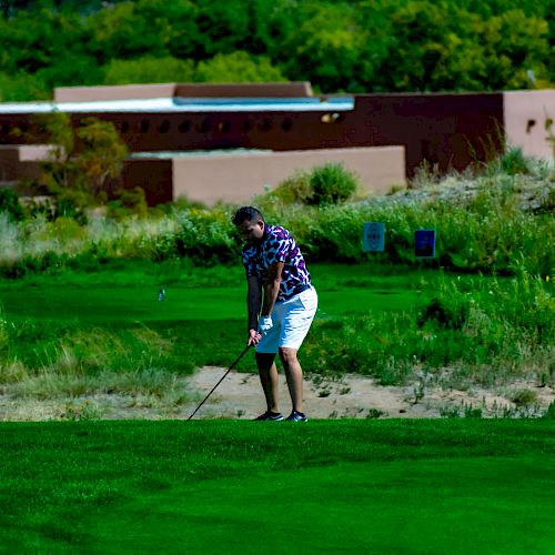 A person is playing golf on a lush green course with buildings and trees in the background on a sunny day.