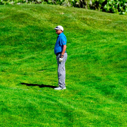 A person dressed in a blue shirt, gray pants, and a white cap stands on a lush green lawn, possibly a golf course, holding a golf club.