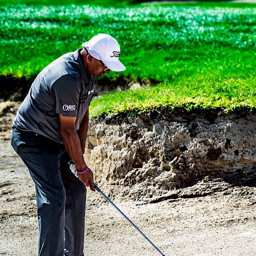 A person in a golf attire is preparing to hit a golf ball from a sand bunker on a golf course, with green grass in the background.