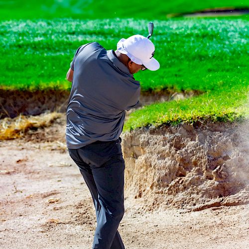 A golfer is hitting a shot from a sand bunker on a sunny day, with vibrant green grass in the background.