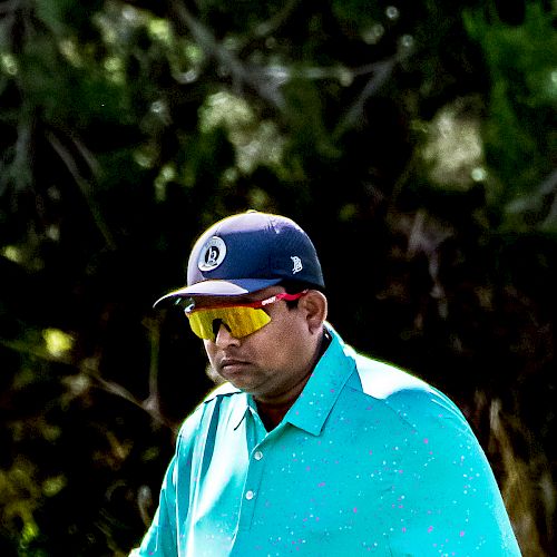 A person wearing a teal polo shirt, black hat, and yellow-tinted glasses is outdoors, with a blurred, leafy background.