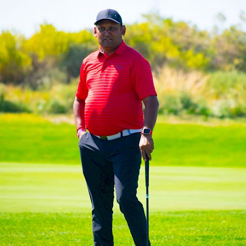 A person in a red shirt and dark pants is standing on a golf course holding a golf club, with green grass and trees in the background.