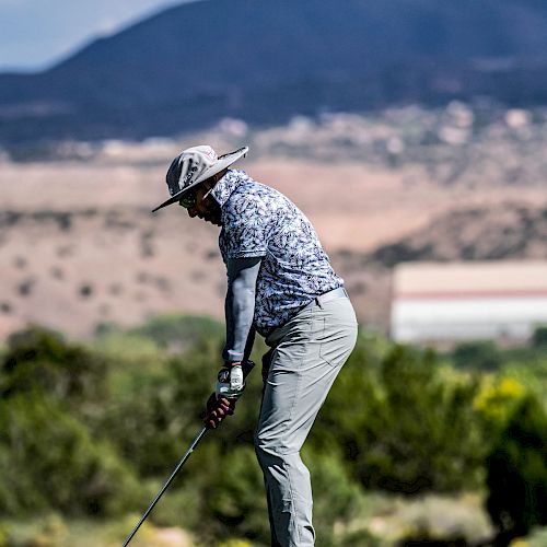 A person is playing golf on a lush green course with a scenic mountainous background, wearing a hat and preparing to swing a club.