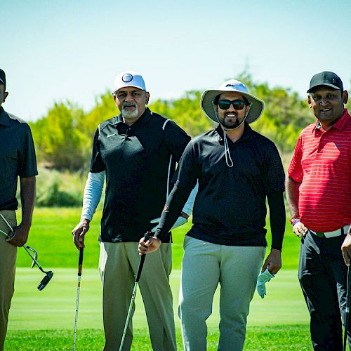 Four men are standing on a golf course, each holding a golf club, and posing for the photo. They are dressed in casual golfing attire and smiling.