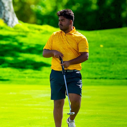 A person wearing a yellow shirt and shorts is standing on a golf course, holding a golf club, with lush green foliage in the background.