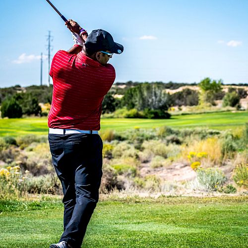 A person wearing a red shirt, black pants, and a hat is swinging a golf club on a golf course with greenery and a clear blue sky in the background.