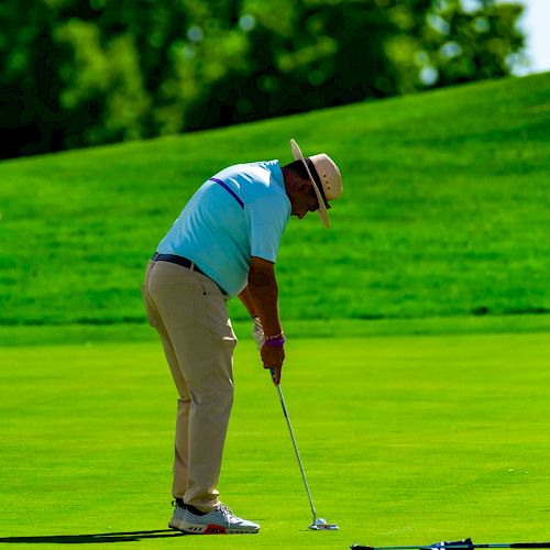 A person wearing a hat is playing golf, focusing on putting on a green course with trees in the background, under a clear, sunny sky.