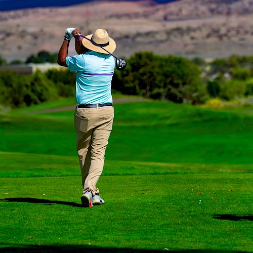 A person in a hat and light clothing is swinging a golf club on a lush green golf course with trees and mountains in the background.