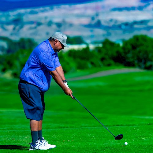 A person is golfing on a lush green course, preparing to swing with a club against a scenic backdrop of hills and clear blue sky.