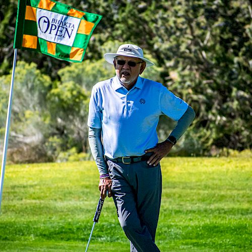 A person is standing on a golf course, leaning on a golf club near a flag that says 