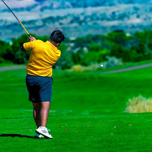 A golfer in a yellow shirt and dark shorts is swinging a club on a vibrant green golf course with scenic hills in the background.