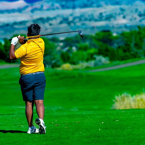 A person in a yellow shirt and shorts is playing golf on a lush green golf course with a mountainous landscape in the background.