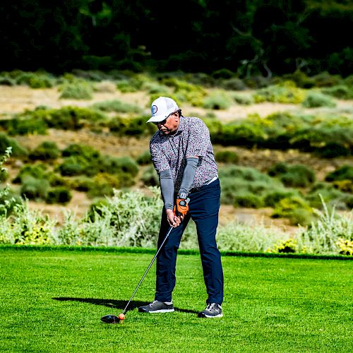 A person wearing a hat and golfing attire is swinging a golf club on a green golf course with a scenic background of trees and hills.