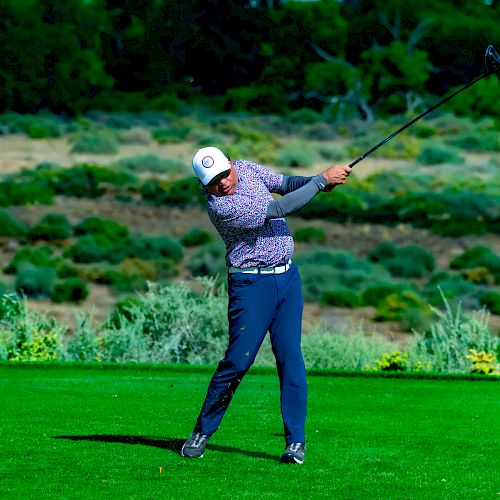 A golfer is taking a swing on a lush green course with a scenic background of trees and hills.