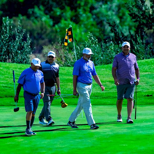 Four people are walking on a golf course near a flagstick, holding golf clubs, wearing casual golf attire and hats, under a sunny sky.