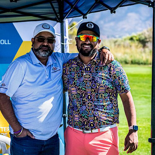 Two men are posing together under a tent at a golf course, with a promotional banner behind them.