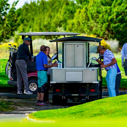 Several people at a golf course gather around a mobile refreshment cart set between golf carts, under a sunny sky with lush greenery in the background.