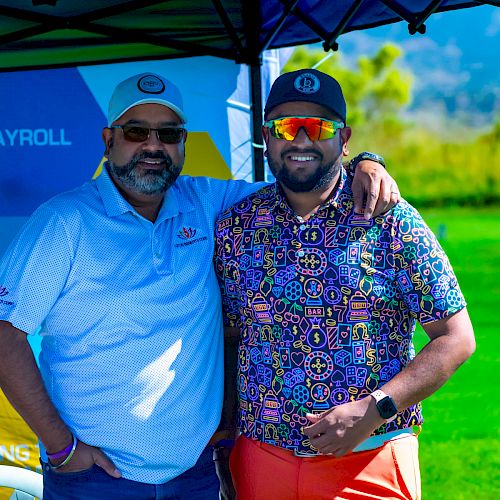 Two men in hats posing under a canopy at a golf event, with a bright outdoor setting.