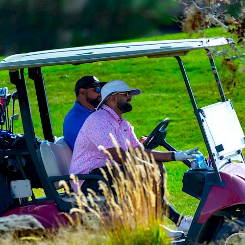 Two men are riding a golf cart on a grassy golf course, with golf bags and equipment visible at the back of the cart, ending the sentence.