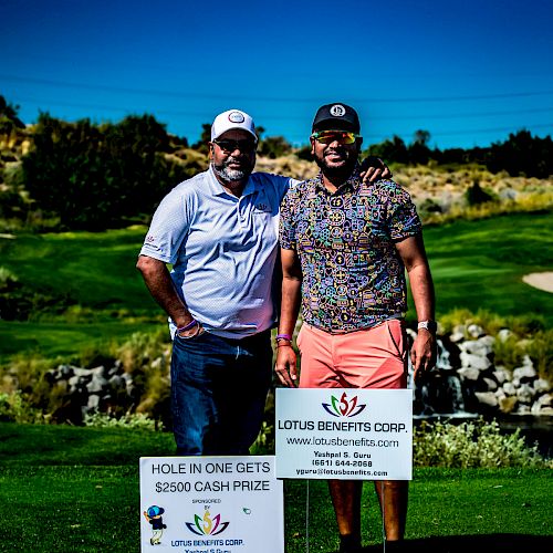 Two men are standing on a golf course, smiling for the photo. They are next to signs promoting a cash prize for a hole in one.