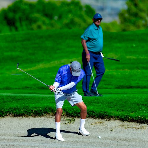 Two men are on a golf course, with one swinging to hit a ball out of a sand trap while the other looks on.