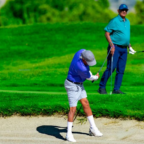 Two people are on a golf course, one is taking a swing in a sand bunker while the other watches with golf clubs in hand, on a sunny day.