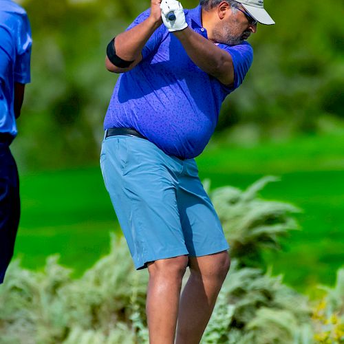 A person wearing a cap, blue shirt, and blue shorts is swinging a golf club on a green golf course with trees in the background.