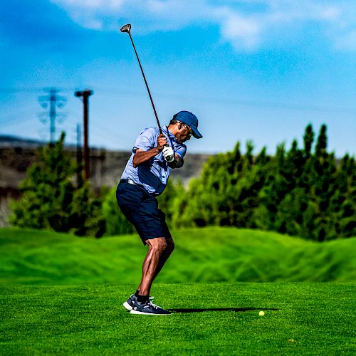 A person is swinging a golf club on a lush green golf course, with trees and a partially cloudy sky in the background. Play is in progress.